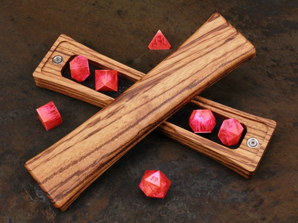 Zebrawood curved dice box with foam lining, opened to reveal pink polyhedral dice. Resting on a weathered metal table. Handcrafted and perfect for gamers and collectors.